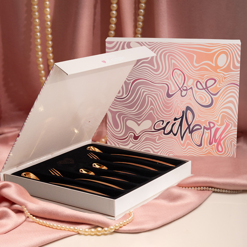 Eine geöffnete weiße Schachtel mit goldenen Love Cutlery-Löffeln liegt auf rosafarbenem Satin 